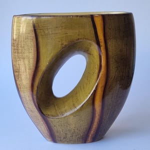 Puede incluir: Un jarrón de cerámica con acabado brillante que presenta una abertura ovalada central. La superficie muestra un patrón texturizado y moteado en tonos verde oliva y marrón oscuro, con vetas verticales de color marrón oscuro en los lados.