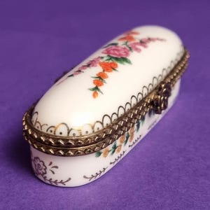 Peut inclure: Boîte à pilules ovale en porcelaine blanche avec couvercle à charnière, ornée d'un motif floral rose, orange et vert. Elle présente une garniture et un fermoir en métal doré, sur fond violet.