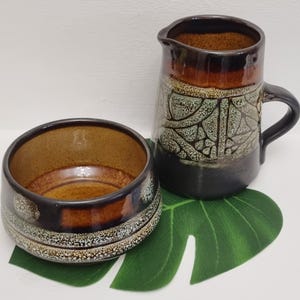 May include: A ceramic bowl and jug set. The bowl is round with a dark brown exterior and a lighter brown interior. The jug has a handle and a spout, with a dark brown top and a black base. The jug has a green and white pattern.