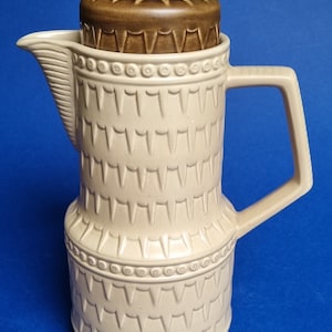 May include: A beige ceramic coffee pot with a brown lid and a geometric pattern. The pot has a handle and a spout.