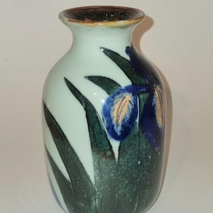 May include: A ceramic vase with a pale light blue base colour, featuring a hand-painted design of dark green leaves and deep blue iris flowers with yellow accents. The vase has a rounded body, a narrow neck, and a dark brown rim at the opening.