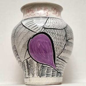 May include: A ceramic vase with a rounded body and a flared rim. The surface features a white base with black hand-drawn patterns including lines, dots, and dashes. A prominent, solid purple teardrop-shaped design is centred on the front.