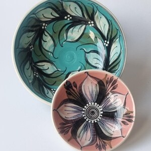 May include: A large turquoise ceramic bowl featuring a black painted leaf pattern with white dots, paired with a smaller pink ceramic bowl decorated with a central black and white floral design featuring a spiral centre and pointed petals.