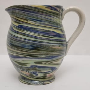 May include: A ceramic jug with a cream-coloured handle. The body of the jug features a swirling pattern of blue, green, yellow, and pink hues. The jug has a rounded base and a spout for pouring.