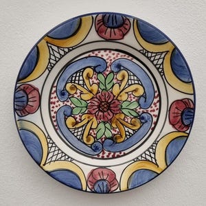 May include: A ceramic plate with a blue rim and a detailed floral pattern. The plate showcases a central floral design in red, yellow, and green, encircled by blue and yellow scrollwork. The background of the plate is white.