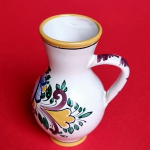 May include: A small white ceramic jug with a yellow rim and a handle. The jug is decorated with a floral pattern in shades of purple, yellow, and green.