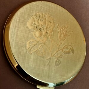 May include: A gold-toned compact mirror with a floral design. The mirror has a raised, engraved design of a rose with leaves and stems.