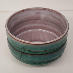 May include: A small, round ceramic bowl with a green and brown glaze. The bowl has a layered design, with the green glaze on the outside and the brown glaze on the inside.