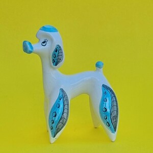 May include: A white ceramic poodle figurine featuring blue accents on the head, nose, and tail. The poodle has painted grey and blue leaf-like patterns on its ears and legs, with black painted eyes and eyelashes on a smooth, glossy surface.