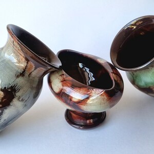 May include: Three ceramic vessels with a glossy finish, featuring a blend of dark brown and muted green glaze. The set includes two vases with flared rims and one central pedestal bowl, all displaying a marbled, reactive colour pattern.