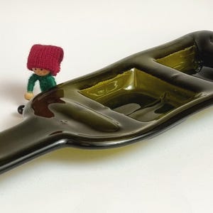 Op de afbeelding: Een donkergroene serveerschaal van gerecycled glas, gemaakt van een wijnfles. De schaal heeft drie compartimenten en een flesvormig ontwerp. Een klein beeldje met een rode hoed en groen shirt staat naast de schaal.