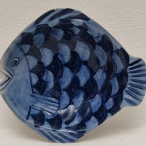 Op de afbeelding: Een keramische, visvormige schaal in blauw en wit. De vis heeft een lachende mond, een oog en een schubbenpatroon. De schaal wordt waarschijnlijk gebruikt om te serveren of te decoreren.