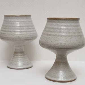 Peut inclure: Deux gobelets en céramique faits à la main. Les gobelets sont gris clair avec une texture mouchetée et un bord marron. La base est en forme de piédestal évasé et le corps a une forme bulbeuse. Idéal pour les boissons.