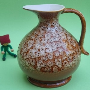 Op de afbeelding: Een bruin-witte keramische kan met een handvat en een tuit. De kan heeft een textuur en een ronde basis. Een kleine pop in een groene outfit en een rode hoed staat op de achtergrond.