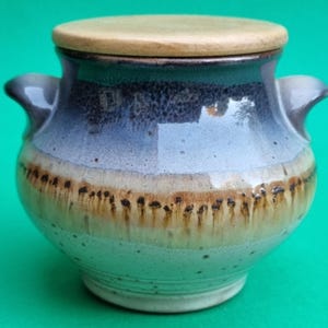 Op de afbeelding: Een handgemaakte keramische pot met een houten deksel. De pot heeft een kleurovergang van blauw, bruin en beige, met donkere accenten. Hij heeft twee kleine handvatten aan weerszijden. Het deksel heeft een lichte, natuurlijke houtkleur.