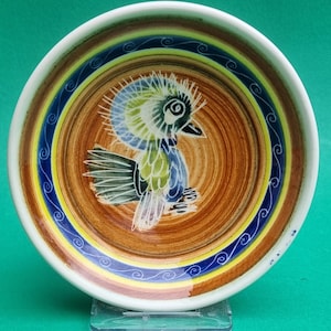 Peut inclure: Un bol en céramique avec un motif d'oiseau peint à la main. L'oiseau est représenté dans des tons de bleu, vert et blanc sur un fond marron. Le bord présente des anneaux concentriques de couleurs, dont le bleu, le jaune et le blanc, avec un motif tourbillonnant décoratif.