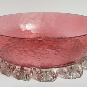 May include: A vintage pink glass bowl with a textured surface and a clear, scalloped base. The bowl's interior has a smooth, rounded shape, and the exterior has a textured, hammered appearance. This decorative bowl is ideal for serving or display.