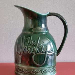 May include: A dark green ceramic jug with a glossy finish, featuring the embossed text The Whisky Shop in a cursive script. The base is decorated with a raised Celtic knot pattern, and the vessel includes a curved handle and a pouring spout.