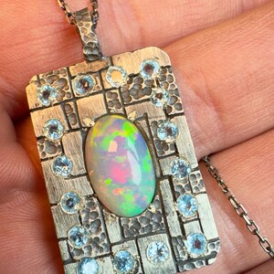 May include: A silver pendant necklace featuring a rectangular pendant with an oval opal gemstone at the center. The pendant is adorned with small, round, light blue gemstones and has a textured, hammered design. The necklace chain is also silver.