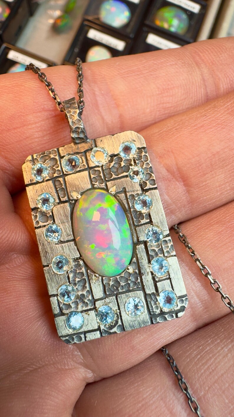 May include: A silver pendant necklace featuring a rectangular pendant with an oval opal gemstone at the center. The pendant is adorned with small, round, light blue gemstones and has a textured, hammered design. The necklace chain is also silver.