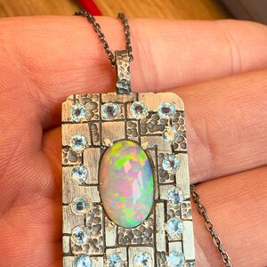 May include: A silver pendant necklace with an oval opal center stone. The pendant is rectangular with a textured border and small blue gemstones. The opal displays a rainbow of colors. The chain is a delicate silver.