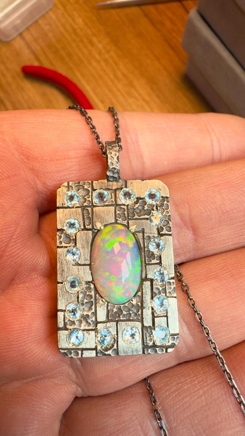 May include: A silver pendant necklace with an oval opal center stone. The pendant is rectangular with a textured border and small blue gemstones. The opal displays a rainbow of colors. The chain is a delicate silver.