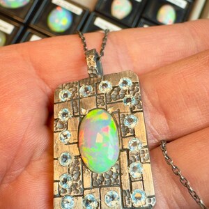 May include: Silver pendant necklace featuring an oval opal gemstone with iridescent colors. The pendant is rectangular, with a textured design and small blue gemstones. The necklace chain is also silver.