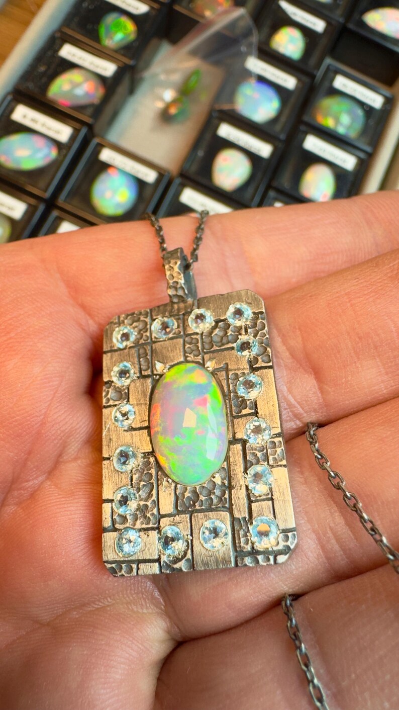 May include: Silver pendant necklace featuring an oval opal gemstone with iridescent colors. The pendant is rectangular, with a textured design and small blue gemstones. The necklace chain is also silver.
