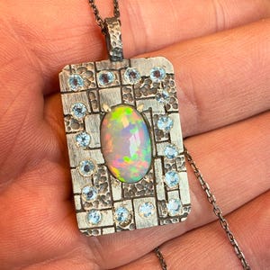 May include: A silver pendant necklace with an oval opal gemstone at the center, surrounded by small blue gemstones. The pendant has a rectangular shape with a textured surface and is suspended from a delicate chain.