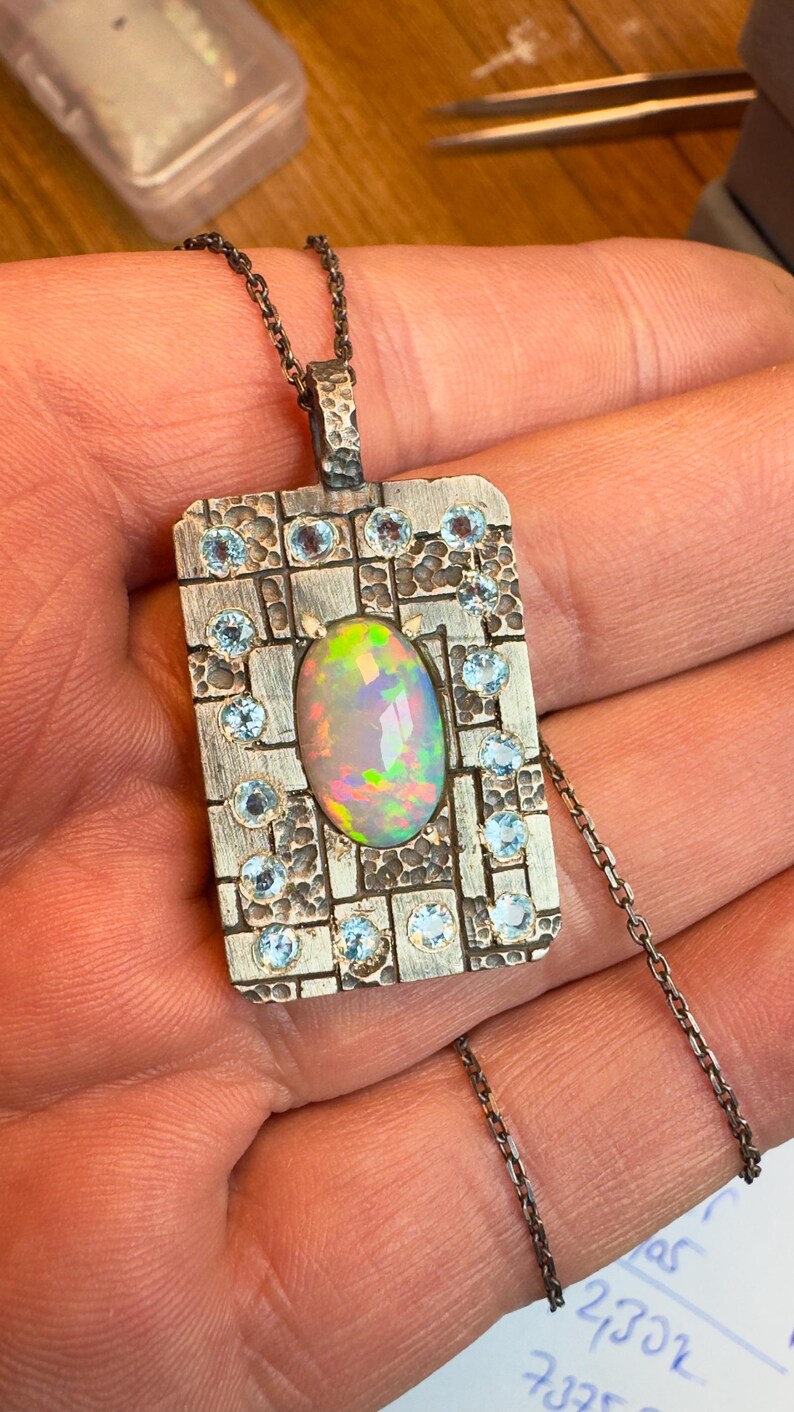 May include: A silver pendant necklace with an oval opal gemstone at the center, surrounded by small blue gemstones. The pendant has a rectangular shape with a textured surface and is suspended from a delicate chain.