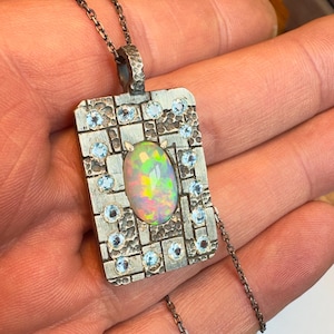 May include: A rectangular silver pendant with an oval opal center stone and small blue gemstones. The pendant is on a silver chain. The opal has a rainbow iridescence. The pendant is held in a hand.