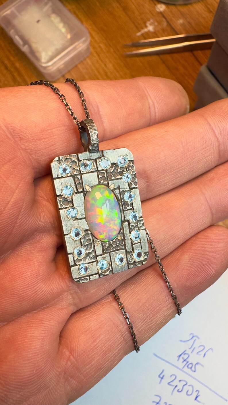 May include: A rectangular silver pendant with an oval opal center stone and small blue gemstones. The pendant is on a silver chain. The opal has a rainbow iridescence. The pendant is held in a hand.