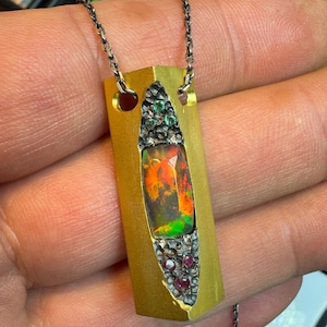 May include: A rectangular pendant necklace with a gold-colored metal frame. The pendant features a central opal stone with vibrant orange, red, and green hues, surrounded by small green and red gemstones. The necklace has a delicate silver chain.