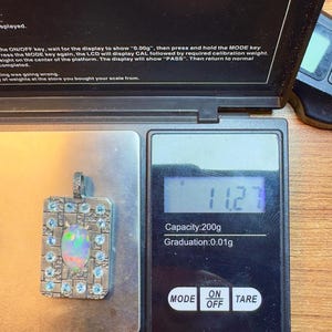 May include: A digital pocket scale with a silver pendant displaying 11.2. The scale has a capacity of 200g and a graduation of 0.01g. The pendant is rectangular with a central opal and blue gemstones.