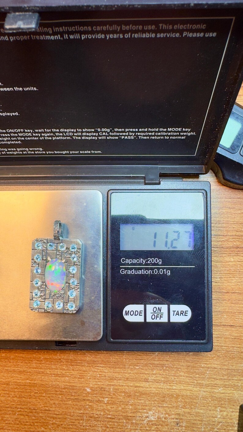 May include: A digital pocket scale with a silver pendant displaying 11.2. The scale has a capacity of 200g and a graduation of 0.01g. The pendant is rectangular with a central opal and blue gemstones.