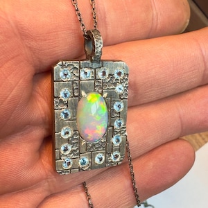 May include: A silver pendant necklace with a rectangular pendant. The pendant features a central oval opal gemstone surrounded by small blue gemstones and a textured silver frame. The necklace chain is also silver.