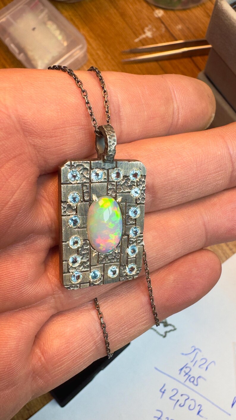 May include: A silver pendant necklace with a rectangular pendant. The pendant features a central oval opal gemstone surrounded by small blue gemstones and a textured silver frame. The necklace chain is also silver.