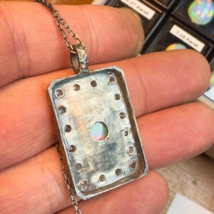 May include: Silver pendant necklace with a rectangular frame, featuring a central opal stone and surrounding clear gemstones. The pendant hangs from a delicate chain. The background shows other opals in small display cases.