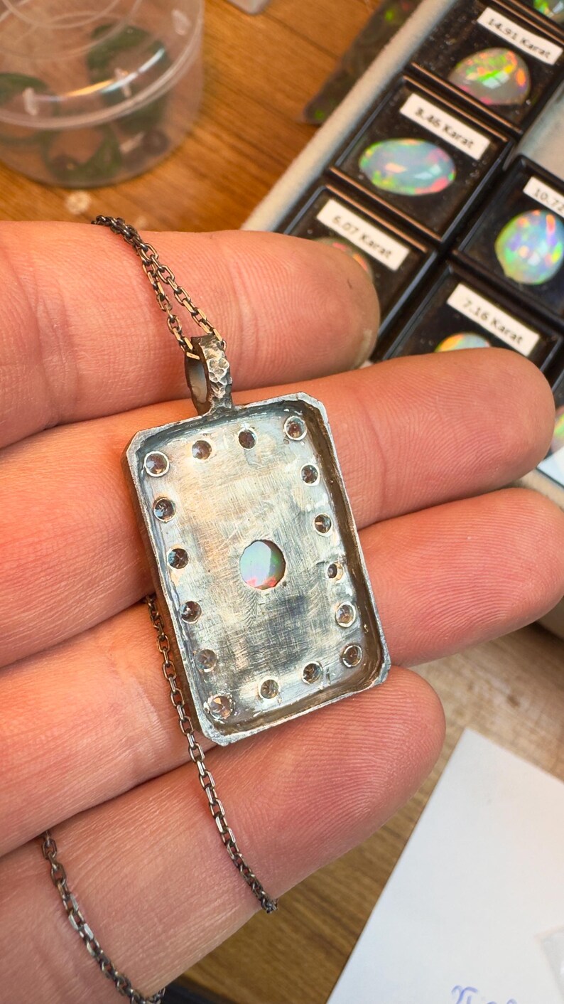 May include: Silver pendant necklace with a rectangular frame, featuring a central opal stone and surrounding clear gemstones. The pendant hangs from a delicate chain. The background shows other opals in small display cases.