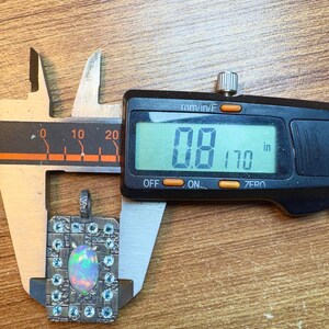 May include: A square pendant with an opal center stone and small blue accent stones is measured with a digital caliper. The digital display reads 0.8 inches. The pendant is set in a silver-colored metal and is on a wooden surface.