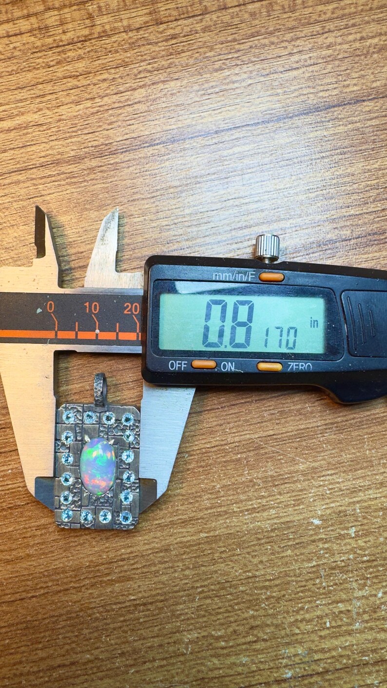 May include: A square pendant with an opal center stone and small blue accent stones is measured with a digital caliper. The digital display reads 0.8 inches. The pendant is set in a silver-colored metal and is on a wooden surface.