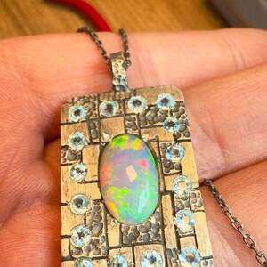 May include: Silver pendant necklace featuring an oval opal gemstone at the center, surrounded by small, round, light blue gemstones. The pendant has a rectangular shape with a textured, grid-like design. The chain is a delicate silver link.