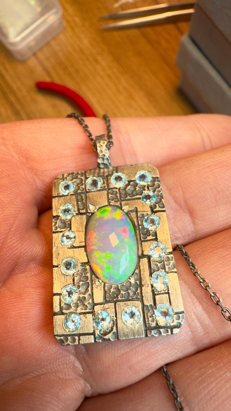 May include: Silver pendant necklace featuring an oval opal gemstone at the center, surrounded by small, round, light blue gemstones. The pendant has a rectangular shape with a textured, grid-like design. The chain is a delicate silver link.