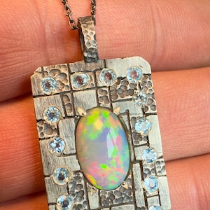 May include: A rectangular pendant necklace with a textured silver finish. The pendant features an oval opal gemstone in the center, surrounded by small, round, light blue gemstones. The necklace chain is also silver.