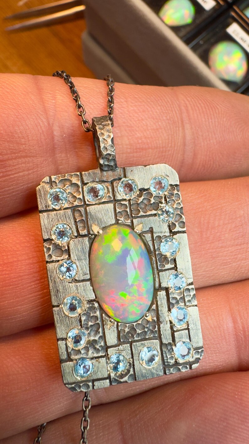 May include: A rectangular pendant necklace with a textured silver finish. The pendant features an oval opal gemstone in the center, surrounded by small, round, light blue gemstones. The necklace chain is also silver.