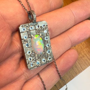 May include: A silver pendant necklace with an oval opal gemstone at the center, surrounded by small blue gemstones. The pendant has a rectangular shape with a textured surface and is suspended from a delicate chain.