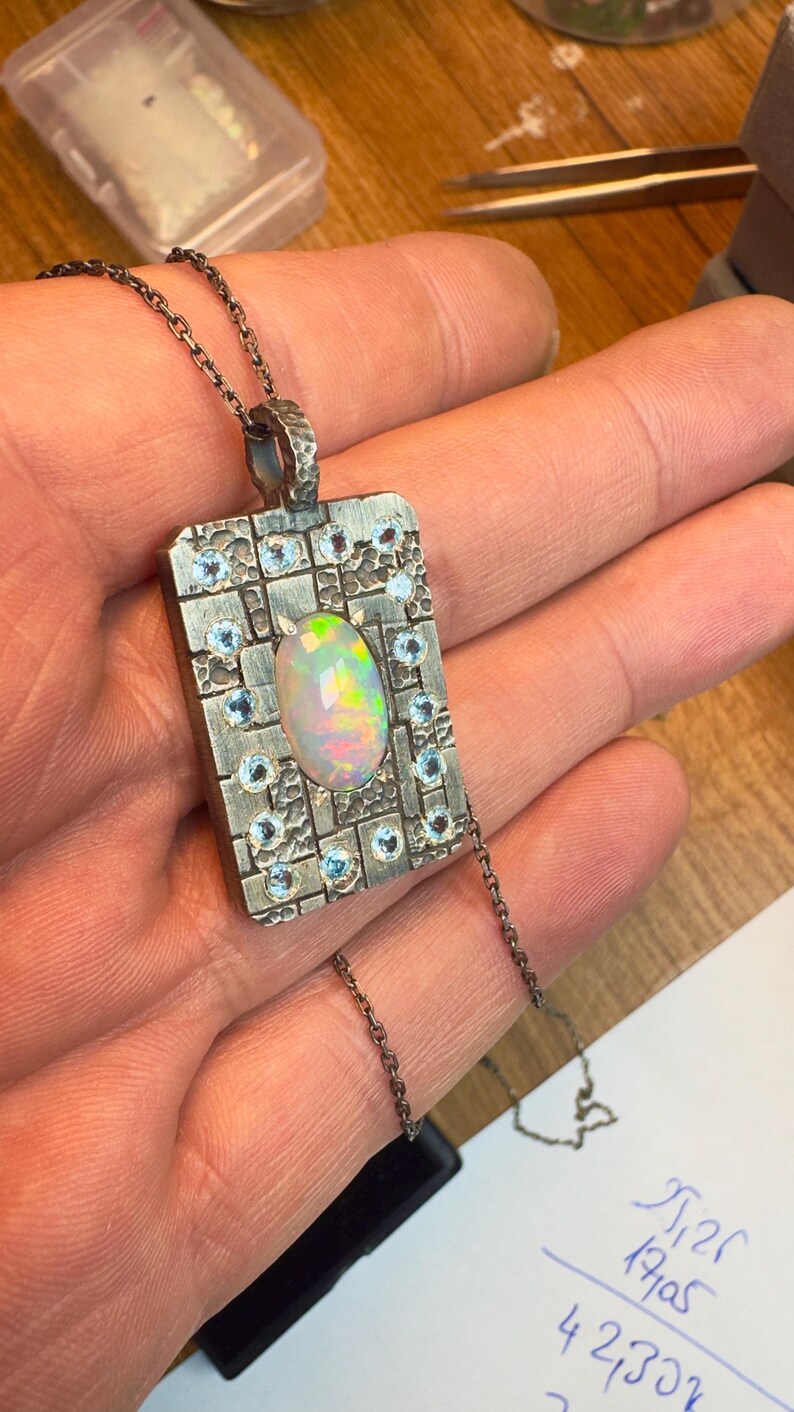 May include: A silver pendant necklace with an oval opal gemstone at the center, surrounded by small blue gemstones. The pendant has a rectangular shape with a textured surface and is suspended from a delicate chain.