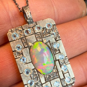 May include: A silver pendant necklace with an oval opal gemstone at the center, surrounded by small, round, light blue gemstones. The pendant has a textured, rectangular design with a chain.