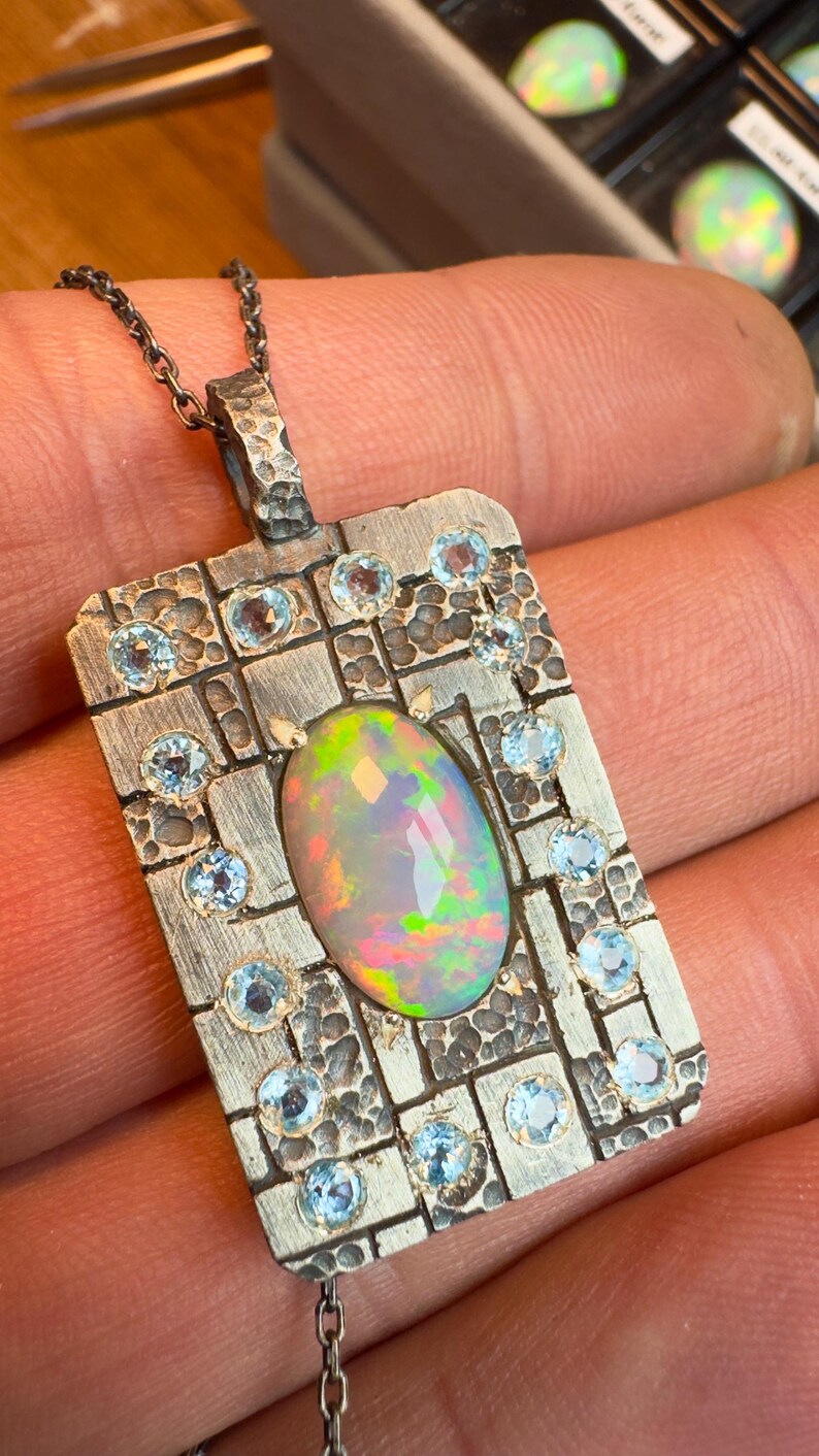 May include: A silver pendant necklace with an oval opal gemstone at the center, surrounded by small, round, light blue gemstones. The pendant has a textured, rectangular design with a chain.