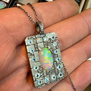May include: A silver pendant necklace featuring a rectangular pendant with an oval opal gemstone at the center. The pendant is adorned with small, round, light blue gemstones and a textured design. The necklace chain is also silver.